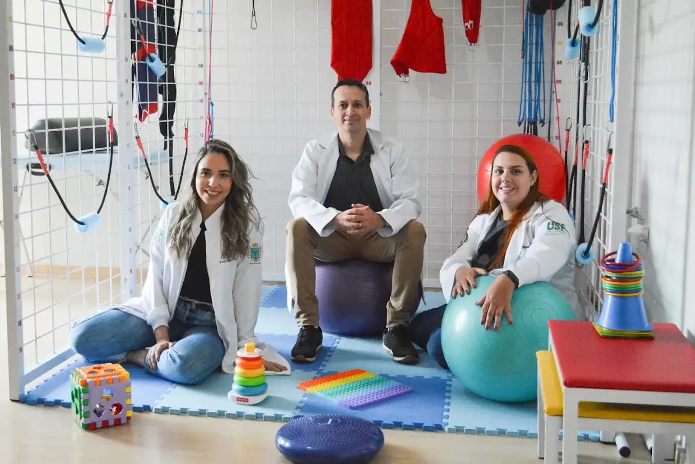 Três profissionais da equipe em uma sala de atendimento com bolas e equipamentos de fisioterapia