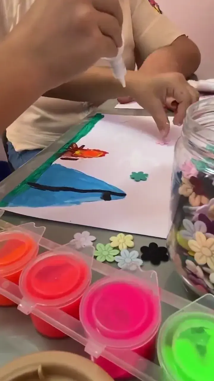 Mãos pintando sobre uma folha de papel, ao lado de potes de tinta colorida e flores de feltro.