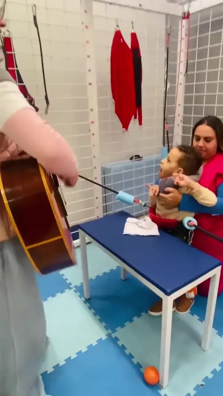 Em uma sala de atendimento com tatame, uma profissional toca violão enquanto outra apoia uma criança em pé.