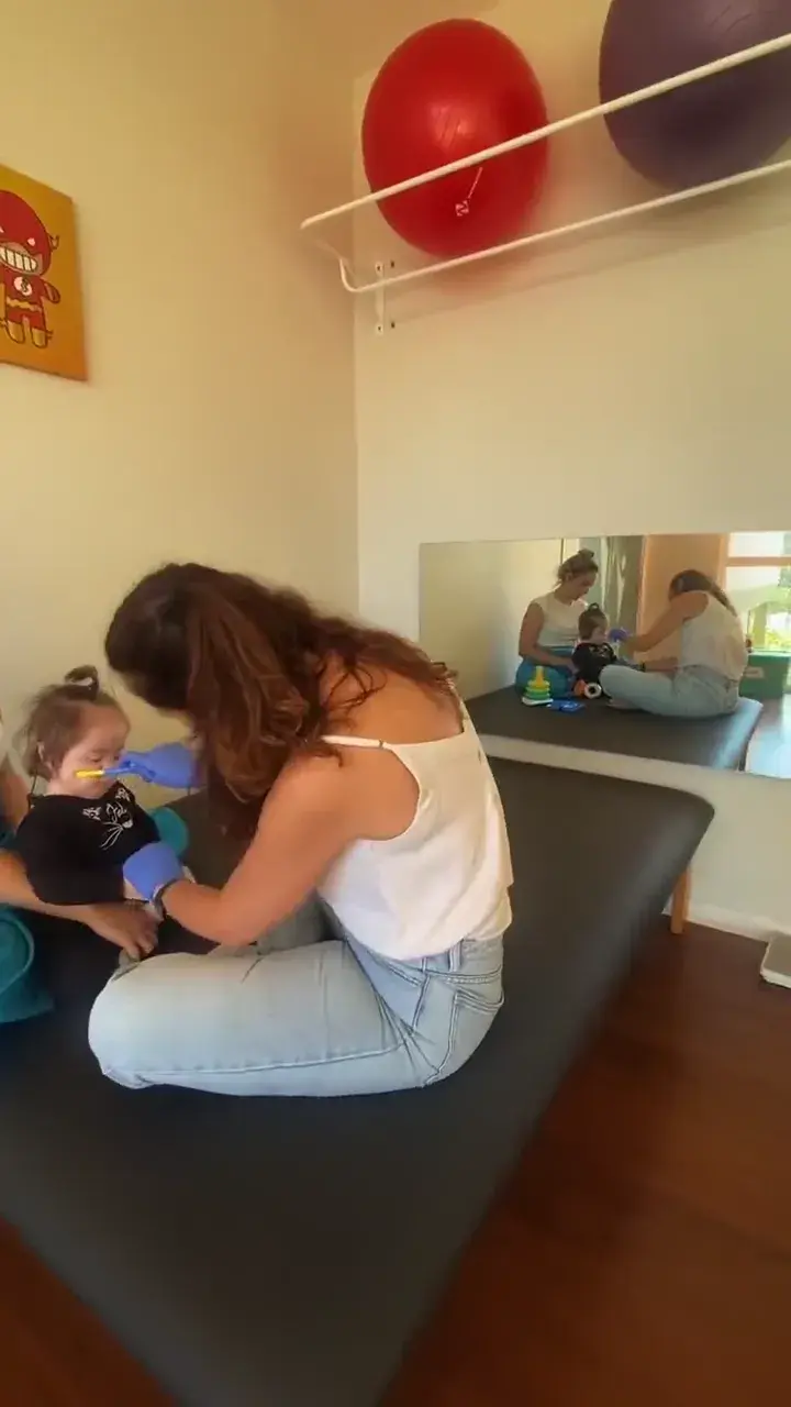 Profissional agachada durante um atendimento com uma criança pequena, diante de um espelho de sala de fisioterapia.